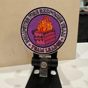 Puede incluir: Un parche bordado de color rosa y blanco con un incendio en un contenedor de basura y el texto "Dumpster Fire Response Team Team Leader".