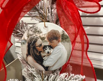 Peut inclure: Une décoration de Noël ronde en céramique avec une photo d'un couple et d'un chien. L'ornement est suspendu par une ficelle dorée et orné d'un ruban rouge et de feuillage givré. Le mot "ONOR" est visible sur l'ornement.