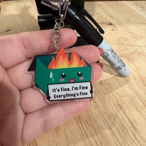 May include: A clear acrylic keyring featuring a green dumpster on fire with a smiling face. The keyring has the text "It's Fine, I'm Fine Everything's Fine" in a white box. A silver keyring and clasp are attached.