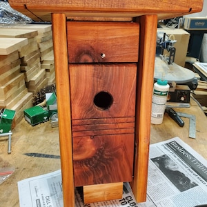 May include: A handcrafted wooden birdhouse with a natural finish. The birdhouse features a rectangular design with a round entry hole and a small perch. The wood grain is visible, and the birdhouse has a small roof.