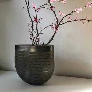 Könnte beinhalten: Eine dunkelbronze-farbene, strukturierte Vase mit abgerundeter Form. Die Vase ist mit Zweigen mit rosa Blüten gefüllt. Die Vase hat ein einzigartiges Muster aus horizontalen und vertikalen Linien, das ein gitterartiges Design erzeugt.