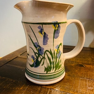 May include: A white ceramic pitcher with a handle, decorated with a blue and green floral pattern. The pitcher has a wide mouth and a rounded base.