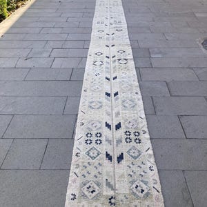 May include: A long, rectangular, off-white rug with a repeating geometric pattern in shades of blue, gray, and beige. The rug has a fringed edge and is placed on a gray stone surface. The design includes diamond and floral motifs.