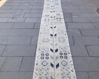 May include: A long, rectangular, off-white rug with a repeating geometric pattern in shades of blue, gray, and beige. The rug has a fringed edge and is placed on a gray stone surface. The design includes diamond and floral motifs.