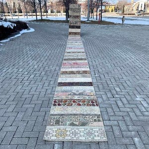 May include: A long, narrow rug with a patchwork design. The rug is made up of different coloured strips of fabric, each with a unique pattern. The rug is laid out on a grey brick walkway.