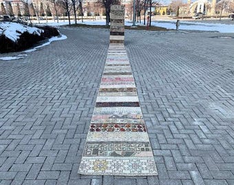 May include: A long, narrow rug with a patchwork design. The rug is made up of different colored strips of fabric, each with a unique pattern. The rug is laid out on a gray brick walkway.