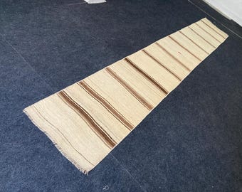 Puede incluir: Una alfombra larga y rectangular con fondo crema y rayas marrones espaciadas. La alfombra tiene un borde con flecos y está colocada sobre una superficie azul oscuro. Esta alfombra es adecuada para un pasillo o entrada. La longitud de la alfombra es de unos 200 cm.