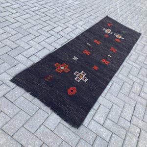 Peut inclure: Un tapis rectangulaire gris foncé avec un motif géométrique rouge, blanc et orange. Le tapis a une bordure effilochée et est posé sur une surface en briques grises. Le motif présente des motifs floraux répétés.