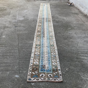 May include: A long, narrow rug with a white background and a blue, brown, and green geometric pattern. The rug has a fringe on both ends.