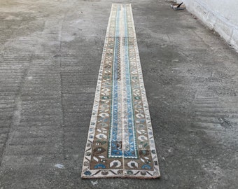 May include: A long, narrow rug with a white background and a blue, brown, and green geometric pattern. The rug has a fringe on both ends.