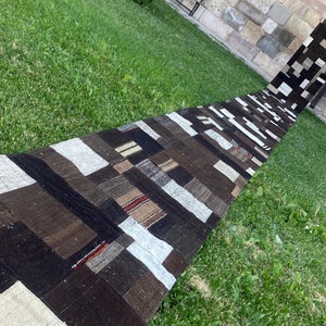 May include: A long, narrow rug made of patchwork fabric in shades of brown, white, and grey. The rug is laid out on a grassy lawn.
