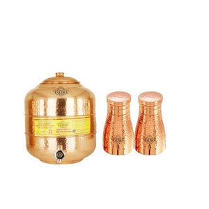 May include: A large hammered copper water dispenser with a lid, a spigot, and a yellow label reading Taluka, accompanied by two matching hammered copper tumblers. Each piece features the Taluka brand logo and a textured, dimpled surface finish.