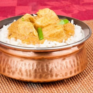 May include: A hammered copper bowl with a silver-toned rim containing a serving of white rice topped with a golden-brown chicken curry and bright green chili pepper slices. The bowl has a rounded base and a wide opening for serving food.