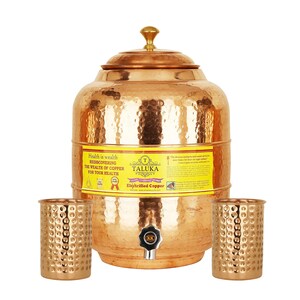 May include: A hammered copper water dispenser with a lid and a spigot, accompanied by two matching hammered copper tumblers. A yellow label on the dispenser features the text Taluka, Electrified Copper, and Health is wealth.