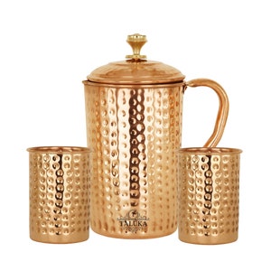 May include: A hammered copper pitcher with a lid and two matching hammered copper tumblers. The pitcher features a gold-toned knob on the lid, a curved handle, and the brand name TALUKA printed in black on the front surface of the vessel.