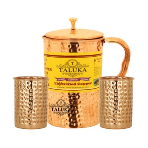 Pode incluir: Uma jarra de cobre com tampa e alça, acompanhada por dois copos de cobre martelado a condizer. A jarra apresenta um rótulo amarelo com o texto TALUKA, Indian Copper Tableware e Electrified Copper impresso na superfície frontal.