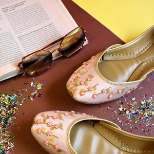 May include: A pair of pink embroidered flat shoes with gold trim and floral embellishments. The shoes are on a brown surface with colorful confetti.