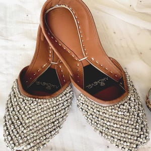 May include: A pair of silver embellished flat shoes with a brown leather insole. The shoes have a pointed toe and are decorated with rows of silver beads. The brand name "ALHOVEYEL" is printed on the insole.
