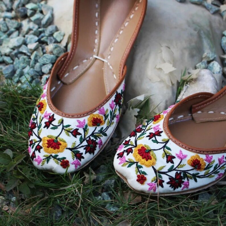 May include: A pair of white embroidered flat shoes with floral designs in shades of pink, yellow, red, and green. The shoes have brown leather soles and a pointed toe.