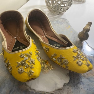 May include: A pair of yellow embroidered flat shoes with gold and white embellishments. The shoes have a pointed toe and a low heel. The brand name "AHOJYEI" is visible on the insole.