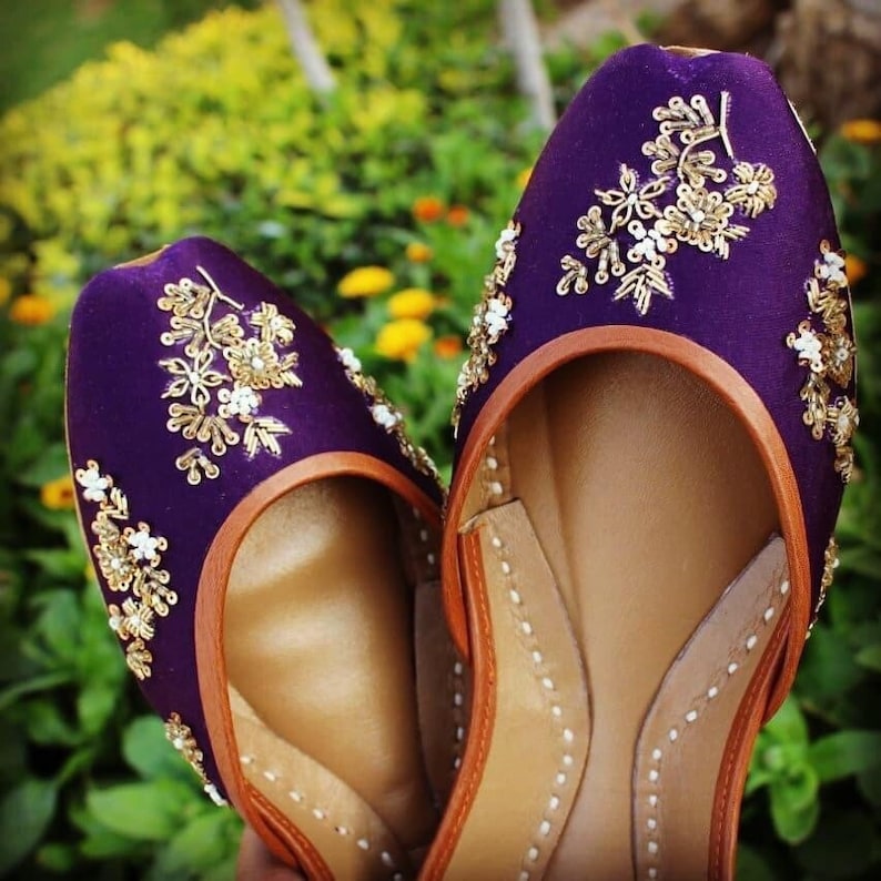 May include: A pair of purple embroidered flat shoes with gold and white embellishments. The shoes have a brown leather insole and a pointed toe.