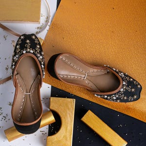 Pode incluir: Um par de sapatos baixos pretos bordados com detalhes em p&eacute;rolas. Os sapatos est&atilde;o sobre uma superf&iacute;cie branca com detalhes dourados e pretos.