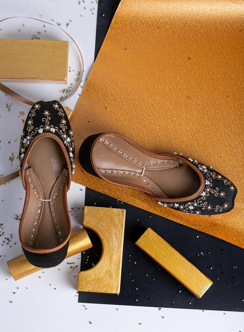 Pode incluir: Um par de sapatos baixos pretos bordados com detalhes em p&eacute;rolas. Os sapatos est&atilde;o sobre uma superf&iacute;cie branca com detalhes dourados e pretos.