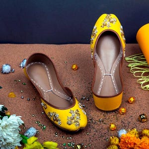 May include: A pair of yellow embroidered flat shoes with gold embellishments. The shoes are on a brown surface with small decorative beads.