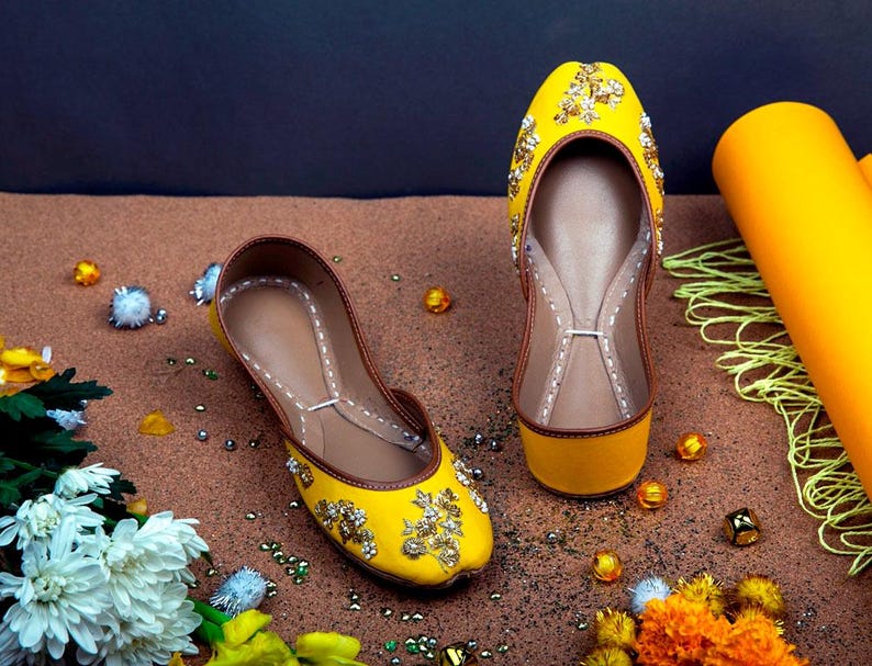 May include: A pair of yellow embroidered flat shoes with gold embellishments. The shoes are on a brown surface with small decorative beads.