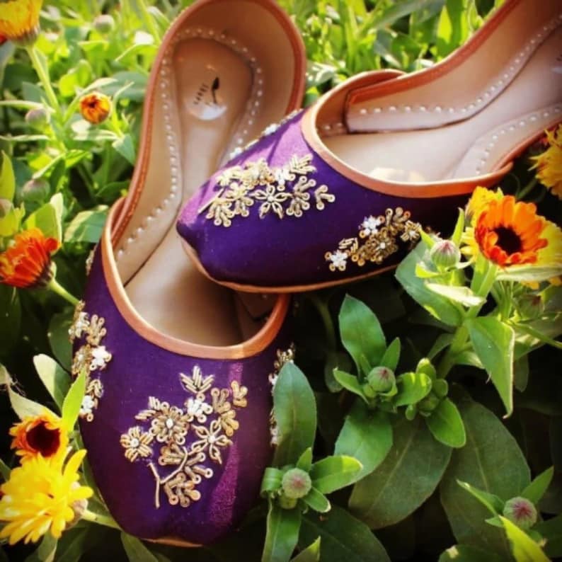 May include: A pair of purple flat shoes with gold and white embroidery. The shoes are sitting on a bed of green leaves and yellow flowers.