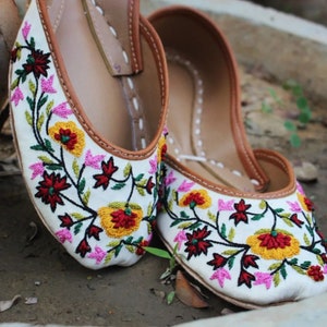 May include: A pair of white embroidered flat shoes with floral designs in shades of pink, yellow, red, and green. The shoes have a brown leather trim around the edges.