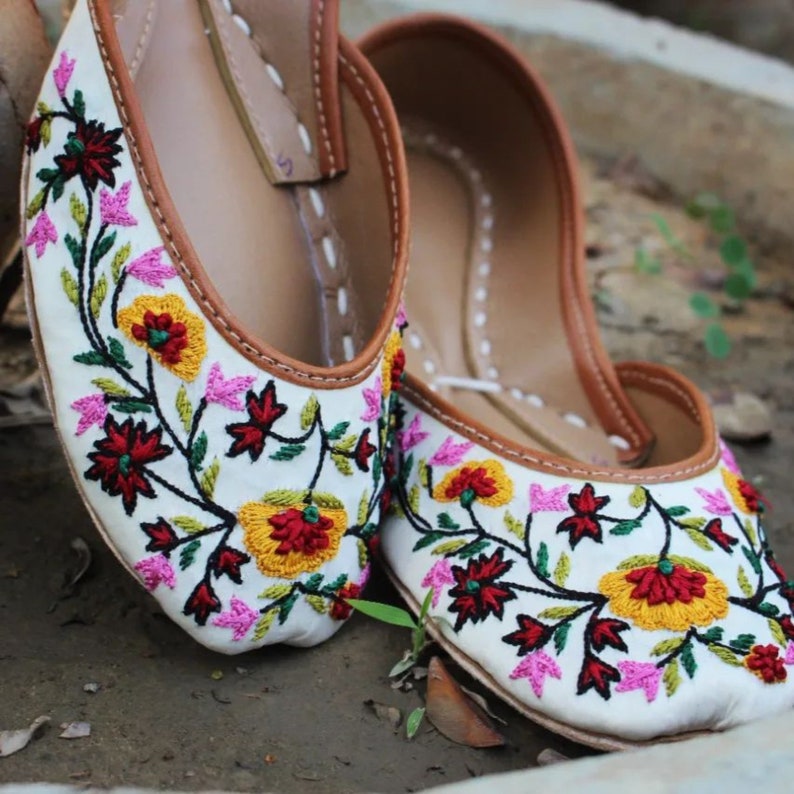 May include: A pair of white embroidered flat shoes with floral designs in shades of pink, yellow, red, and green. The shoes have a brown leather trim around the edges.
