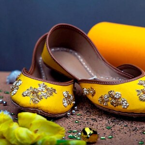 May include: A pair of yellow embroidered flat shoes with gold and white embellishments. The shoes have a brown leather sole and are sitting on a brown surface.