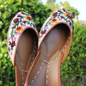 May include: A pair of white embroidered flat shoes with colorful floral designs. The shoes have a brown leather sole and a pointed toe.