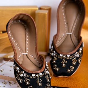 Pode incluir: Um par de sapatos de cetim preto com bordados dourados e brancos. Os sapatos t&ecirc;m um forro de couro castanho e uma ponta pontiaguda.