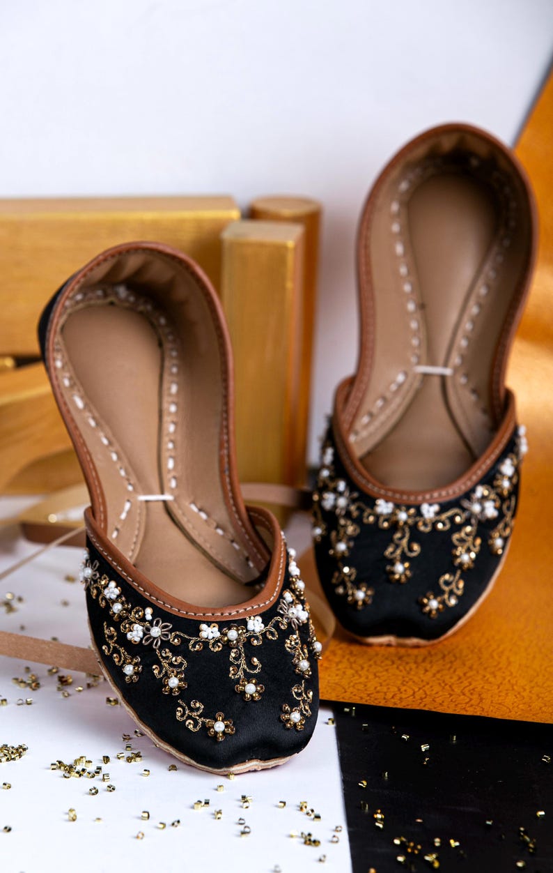Pode incluir: Um par de sapatos de cetim preto com bordados dourados e brancos. Os sapatos t&ecirc;m um forro de couro castanho e uma ponta pontiaguda.
