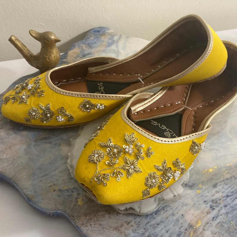 May include: A pair of yellow embroidered flat shoes with gold and white embellishments. The shoes have a pointed toe and a low heel. The brand name "Loveyl" is visible on the insole.