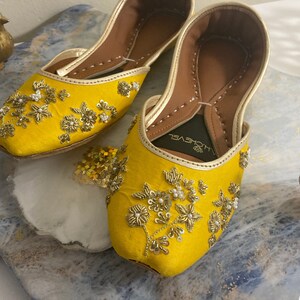 May include: A pair of yellow embroidered flat shoes with gold and white embellishments. The shoes have a pointed toe and a small opening at the top. The brand name "J'CHEVEL" is visible on the inside of one shoe.