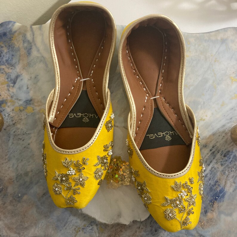 May include: A pair of yellow embroidered flat shoes with gold embellishments. The shoes have a pointed toe and a closed back. The brand name "ALHOVEYEL" is visible on the inside of each shoe.