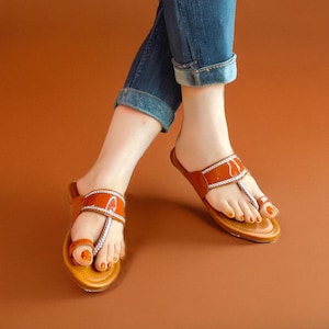 May include: A pair of brown leather sandals with a glossy, orange-brown strap and a silver-colored, woven trim. The sandals have a toe ring design and are worn with blue jeans. The background is a solid, warm brown.
