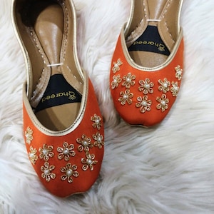 K&ouml;nnte beinhalten: Ein Paar orangefarbene Ballerinas mit goldenen Verzierungen und floralen Stickereien. Die Ballerinas haben eine spitze Zehenpartie und sind mit goldenen und perlenartigen Verzierungen versehen. Die Schuhe werden auf einer wei&szlig;en, flauschigen Oberfl&auml;che pr&auml;sentiert.