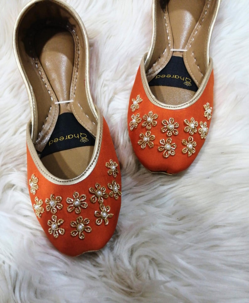 K&ouml;nnte beinhalten: Ein Paar orangefarbene Ballerinas mit goldenen Verzierungen und floralen Stickereien. Die Ballerinas haben eine spitze Zehenpartie und sind mit goldenen und perlenartigen Verzierungen versehen. Die Schuhe werden auf einer wei&szlig;en, flauschigen Oberfl&auml;che pr&auml;sentiert.