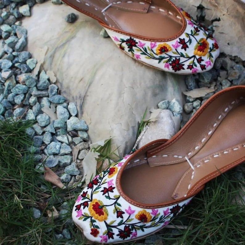 May include: A pair of white embroidered flat shoes with floral designs in shades of pink, yellow, red, and green. The shoes have a brown leather sole and a pointed toe.