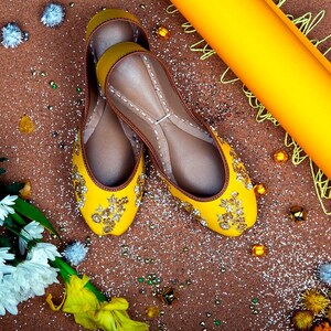 May include: A pair of yellow embroidered flat shoes with gold embellishments. The shoes are on a brown surface with glitter and other decorative items.