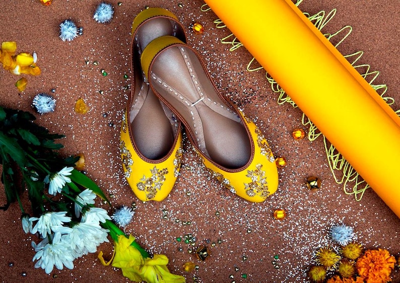 May include: A pair of yellow embroidered flat shoes with gold embellishments. The shoes are on a brown surface with glitter and other decorative items.