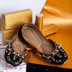 Pode incluir: Um par de sapatos baixos pretos bordados com detalhes dourados e brancos. Os sapatos est&atilde;o sobre um fundo dourado e preto.