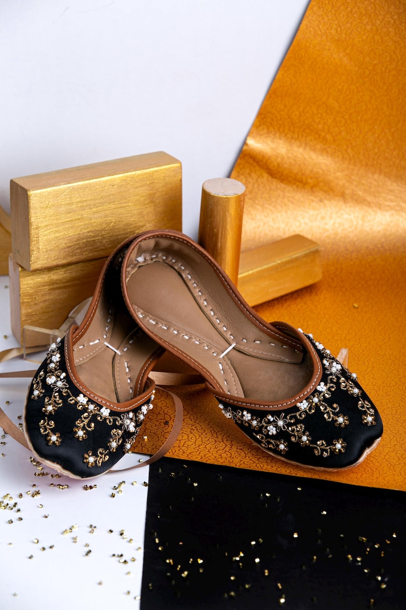 Pode incluir: Um par de sapatos baixos pretos bordados com detalhes dourados e brancos. Os sapatos est&atilde;o sobre um fundo dourado e preto.