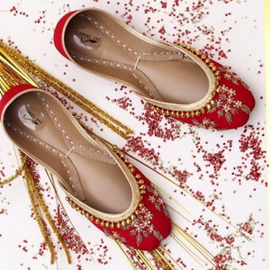 May include: A pair of red embroidered flat shoes with gold embellishments. The shoes are on a white background with red and gold beads scattered around them.