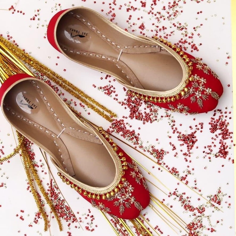 May include: A pair of red embroidered flat shoes with gold embellishments. The shoes are on a white background with red and gold beads scattered around them.