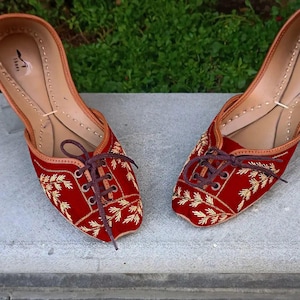 May include: A pair of red velvet shoes with gold embroidery and brown laces. The shoes have a pointed toe and a flat heel.